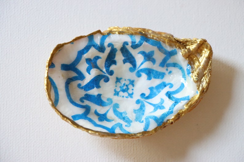 May include: A small, white oyster shell dish with a blue and white patterned design. The shell is painted gold around the edges.
