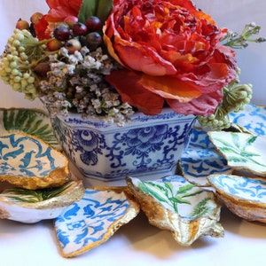 May include: A set of decorative oyster shell dishes with blue and white floral patterns and gold accents. The dishes are arranged around a blue and white porcelain vase with a floral design. The vase is filled with a red flower and green foliage.