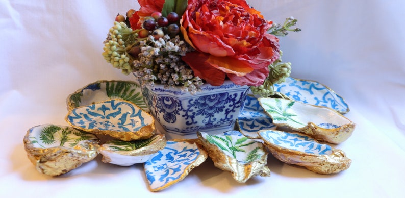 May include: A set of decorative oyster shell dishes with blue and white floral patterns and gold accents. The dishes are arranged around a blue and white porcelain vase with a floral design. The vase is filled with a red flower and green foliage.