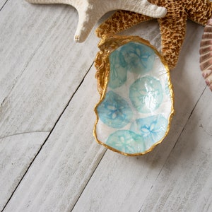 May include: A white ceramic dish with a gold rim and a blue and white sand dollar design. The dish is sitting on a white wooden surface.