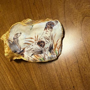 May include: A hand-painted oyster shell with a gold trim, featuring a detailed illustration of a brown and white dog in a grassy field.