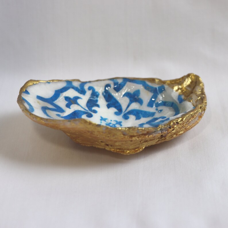 May include: A white ceramic dish with a blue and white floral pattern, with a gold rim. The dish is shaped like a seashell.