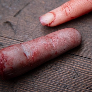 May include: Two severed, bloody fingers on a wooden surface. The fingers are pale and have visible fingerprints. One finger is missing the tip.