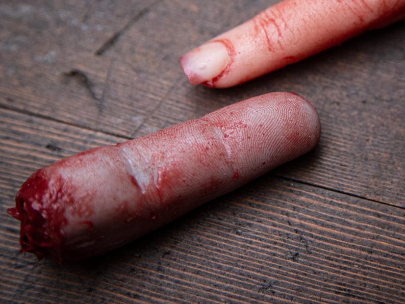 May include: Two severed, bloody fingers on a wooden surface. The fingers are pale and have visible fingerprints. One finger is missing the tip.