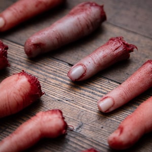 May include: Several severed, bloody fingers are arranged on a wooden surface. The fingers are realistic and appear to be made of a fleshy material.