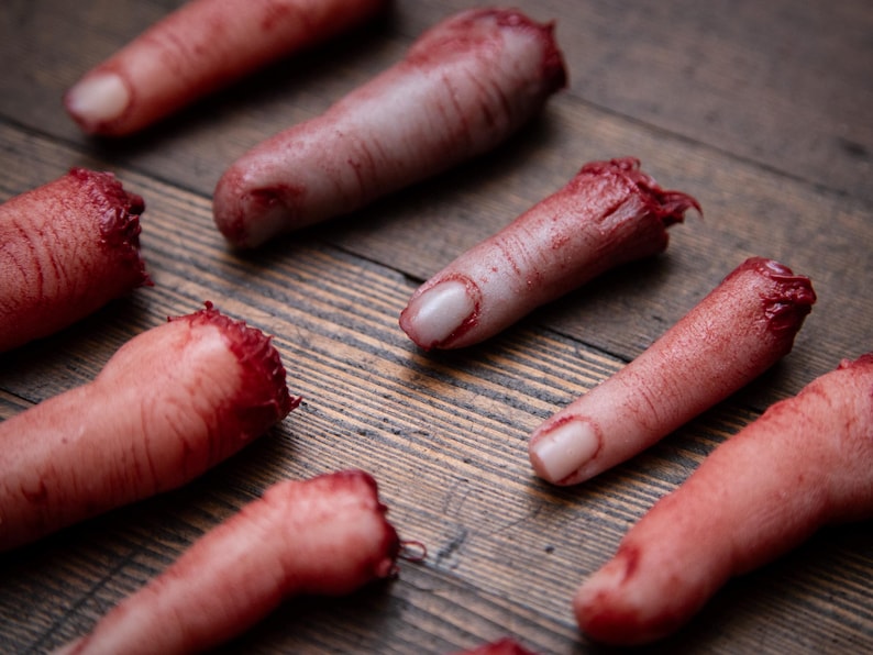 May include: Several severed, bloody fingers are arranged on a wooden surface. The fingers are realistic and appear to be made of a fleshy material.