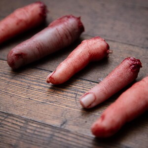 May include: Five severed, bloody fingers on a wooden surface. The fingers are realistic and detailed, with visible blood and flesh.