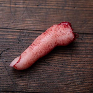 May include: A severed finger prop with realistic detail, covered in fake blood. The finger is lying on a wooden surface.