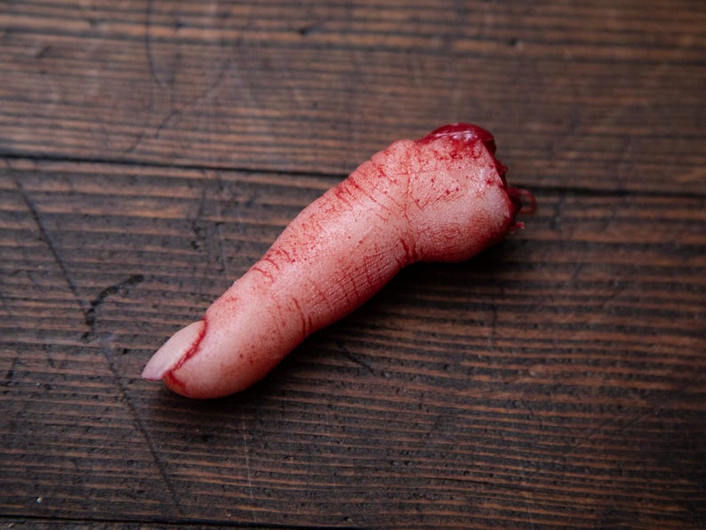 May include: A severed finger prop with realistic detail, covered in fake blood. The finger is lying on a wooden surface.