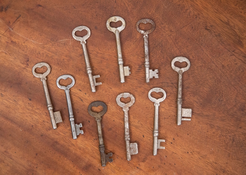 May include: Nine antique skeleton keys with heart-shaped keyholes are arranged on a wooden surface. The keys are made of metal and show signs of age, with a rustic, vintage aesthetic. The keys vary in size and design.