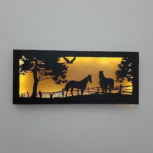 May include: A black metal wall art piece featuring a silhouette of two horses in a field with trees and a fence. The background is a warm yellow colour.