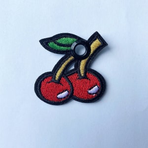 May include: Embroidered patch featuring two red cherries with black outlines, yellow stems, and a green leaf. The cherries have white highlights and a black grommet for attachment. This patch is suitable for clothing or accessories.