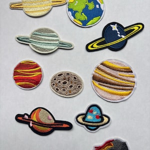 May include: A collection of embroidered planet patches in various designs. Includes patches of Saturn, Earth, and other planets with colorful details. The patches are in shades of yellow, blue, green, red, brown, and orange.
