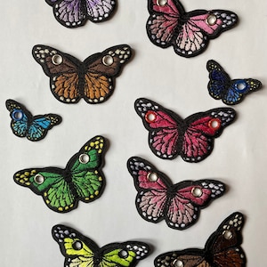 May include: Assortment of ten embroidered butterfly patches in various colors, including purple, pink, orange, blue, green, and yellow. Each patch has two eyelets for attaching.