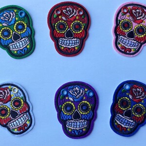 May include: Embroidered skull patches in various colors, including green, red, pink, light blue, purple, and dark blue. Each patch features a floral design with a rose and decorative eyelets. The patches are likely for clothing or craft projects.