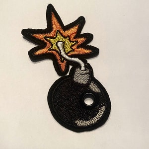 May include: Embroidered patch featuring a black bomb with a silver fuse and a starburst explosion. The starburst has orange, yellow, and black details. The patch is likely for clothing or accessories.