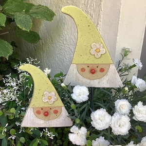 Könnte beinhalten: Zwei Gartenzwerge aus Keramik mit hellgrünen Hüten und weißen Bärten. Jeder Zwerg hat eine Blume auf seinem Hut und eine rote Nase. Die Zwerge stehen in einem Beet mit weißen Blumen und grünem Laub.
