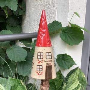 Könnte beinhalten: Ein Keramik-Insektenhotel in Form eines Hauses mit rotem Dach und weißen Wänden. Das Haus hat zwei Fenster und eine Tür. Der Text "Insekten Hotel" steht auf der Vorderseite des Hauses.