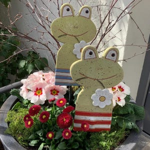Könnte beinhalten: Zwei Gartenstecker aus Keramik in Form von Fröschen mit lächelnden Gesichtern und Blumenakzenten stehen in einem Blumentopf mit rosa und roten Blumen. Die Frösche haben gelbe Körper mit blau-rot gestreiften Details.