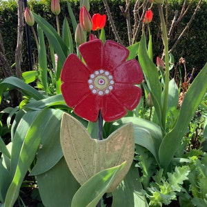 Könnte beinhalten: Rote Keramikblume mit grünen Blättern und einer weißen Mitte. Die Blume ist auf einem schwarzen Metallpfahl und ist von grünen Pflanzen umgeben.