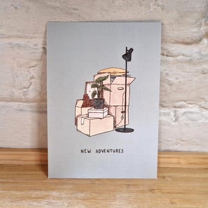 Peut inclure: Carte bleu clair avec une illustration de cartons de déménagement, une plante et une lampe. Les mots "NEW ADVENTURES" sont imprimés en bas. La carte est posée sur une surface en bois contre un mur de briques blanches.