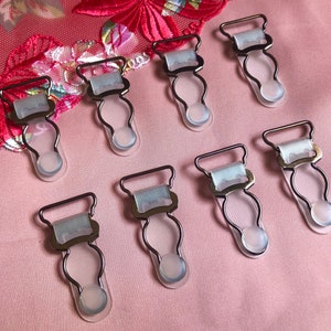 May include: Eight silver-toned metal garment clips with clear plastic straps and white elastic bands. The clips have a rounded shape and are designed to hold fabric together.