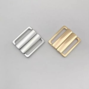 Op de afbeelding: Twee metalen bh-bandjes schuifregelaars. Eén is zilverkleurig, de andere is goudkleurig. Elke schuifregelaar heeft een rechthoekig frame met twee parallelle staven in het midden. De schuifregelaars zijn ontworpen om de lengte van bh-bandjes aan te passen.