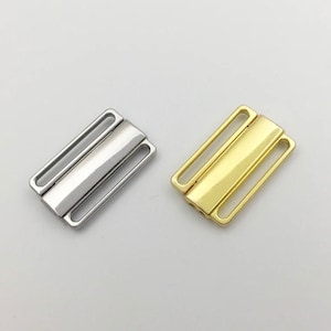 May include: Two metal bra clasps, one silver and one gold. The clasps are rectangular with two loops on each side and a flat bar in the middle.