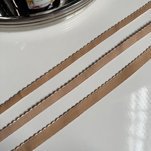 May include: Three pieces of beige satin ribbon with scalloped edges. The ribbon is arranged in a diagonal pattern on a white surface.