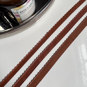 May include: Two brown satin ribbons with scalloped edges. The ribbons are laying flat on a white surface.