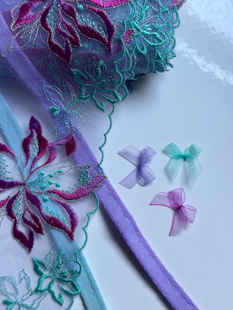 Pu&ograve; includere: Primo piano di un bordino in pizzo viola e blu con ricamo floreale. Tre piccoli fiocchi in tessuto trasparente di colore viola, rosa e turchese sono disposti su una superficie bianca.