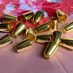 May include: A group of 15 gold-colored metal cord ends. The cord ends are shaped like a cone with a small hole at the top. They are scattered on a pink surface.