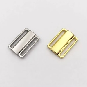 May include: Two metal clasps, one silver and one gold, for attaching straps or fabric. The clasps have a rectangular shape with two parallel slots on each side.