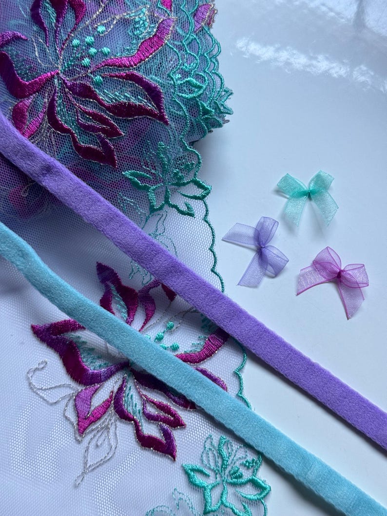 Pu&ograve; includere: Due lunghezze di nastro morbido e vellutato in azzurro e lavanda. Il nastro &egrave; appoggiato su un pezzo di tessuto trasparente con un motivo floreale in tonalit&agrave; turchese, rosa e viola. Ci sono tre piccoli fiocchi in tessuto trasparente in tonalit&agrave; rosa, lavanda e turchese.