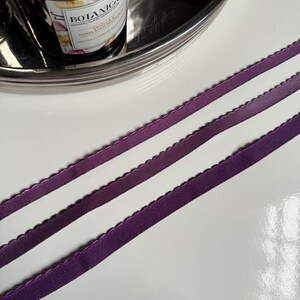 May include: Three purple satin ribbons with scalloped edges. The ribbons are arranged in a row on a white surface.