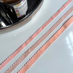 May include: Three pink satin ribbons with scalloped edges. The ribbons are arranged in a diagonal pattern on a white surface.