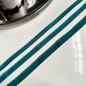 May include: Two teal blue satin ribbons with scalloped edges. The ribbons are lying flat on a white surface.