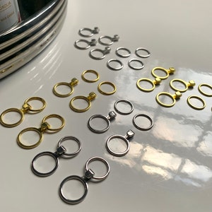 Peut inclure: Assortiment de porte-clés en argent, or et laiton avec un petit charme carré attaché à chaque anneau. Les porte-clés sont disposés en rangées sur une surface blanche.