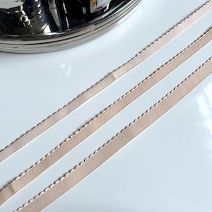 May include: Three pieces of beige elastic ribbon with a scalloped edge. The ribbon is lying flat on a white surface.