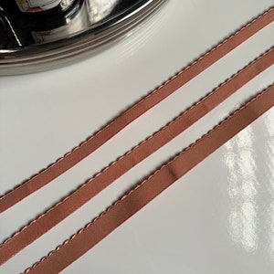 May include: Three strands of brown satin ribbon with scalloped edges. The ribbon is lying flat on a white surface.