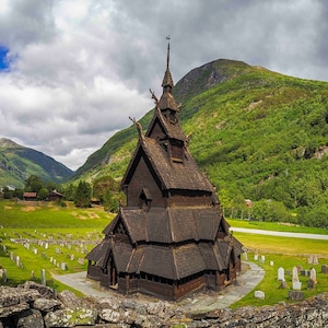 Puede incluir: Una iglesia de madera oscura con intrincadas tallas y una alta aguja se alza en un valle verde. La iglesia está rodeada por un cementerio con lápidas blancas, con montañas al fondo.