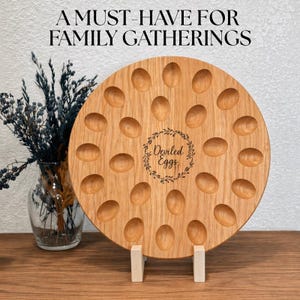 May include: A round wooden deviled egg serving tray with 20 egg-shaped indentations. The tray has a natural wood grain finish and is supported by two white wooden stands. The text "Deviled Eggs" is in the center, surrounded by a wreath. The text "A MUST-HAVE FOR FAMILY GATHERINGS" is at the top.