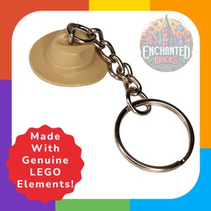 Peut inclure: Un porte-clés en forme de chapeau LEGO beige avec une chaîne et un anneau argentés. Le porte-clés est fabriqué avec de véritables éléments LEGO. Le texte "Made With Genuine LEGO Elements!" est sur un cercle rouge.