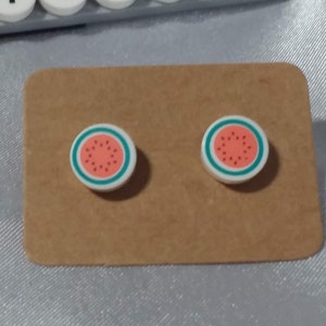 Può includere: Un paio di orecchini a bottone a tema anguria realizzati con mattoncini Lego bianchi. Gli orecchini sono su una carta marrone con il testo "Enchanted Bricks" su una piastra di base Lego grigia.