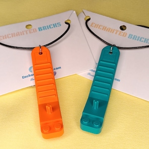 May include: Two necklaces with Lego brick pendants. One pendant is orange and the other is teal. Both necklaces have black cords and are displayed on white cards with the text "Enchanted Bricks" printed on them.