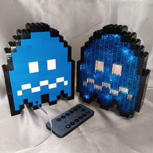 Puede incluir: Dos lámparas pixeladas con forma de fantasma hechas con bloques negros y azules. Una lámpara es azul sólido, la otra brilla con luz azul. Un mando a distancia está delante de las lámparas. Las lámparas están conectadas por un cable.