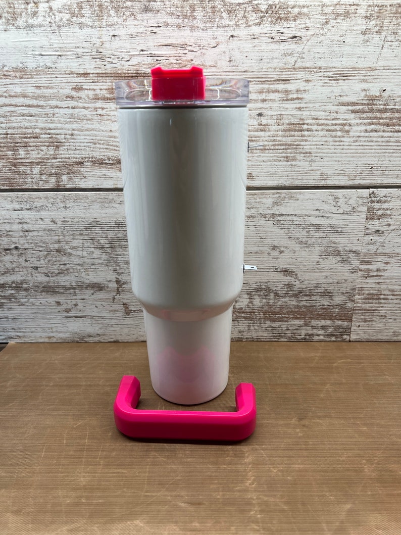 May include: White stainless steel tumbler with a pink lid and a pink handle. The tumbler is sitting on a brown surface.
