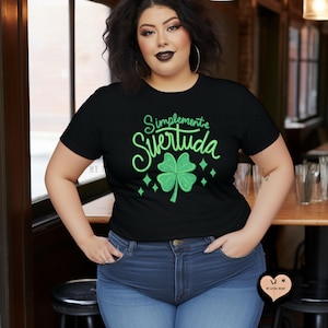May include: Black crew neck t-shirt with the text "Simplemente Suertuda" in green script, featuring a green four-leaf clover and diamond accents. The shirt is paired with blue jeans.