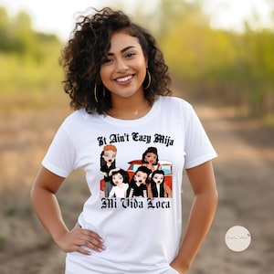 May include: White t-shirt with a graphic of cartoon girls in a red car. The text "It Ain't Eazy Mija Mi Vida Loca" is printed on the shirt.