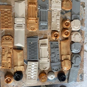 May include: Assortment of small, detailed plastic model pieces in various shades of gray, white, and gold. The pieces have a textured, mechanical look and appear to be parts of a larger model.